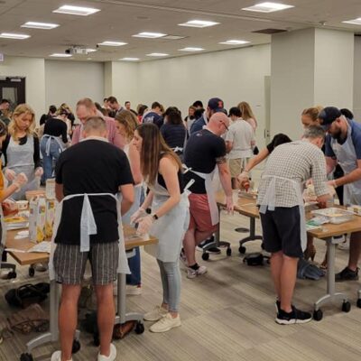 A large group of participants, all wearing aprons, are spread across multiple tables in a spacious room. They are actively preparing food together, working in small teams to assemble meals for donation. The room is well-lit, with several rows of tables and individuals focused on their tasks, contributing to a community service cooking event aimed at making food for those in need. thumbnail