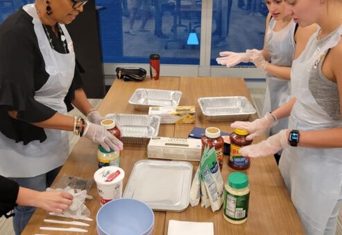 A group of four women, wearing aprons and gloves, are standing around a table during a team building cooking activity. Various ingredients, such as jars of pasta sauce, parmesan cheese, and lasagna trays, are placed on the table, along with mixing bowls and utensils. They are preparing to assemble a dish, with one woman holding a jar of sauce and the others ready to begin. The group appears focused, engaged in the process, and collaborating on the cooking task.