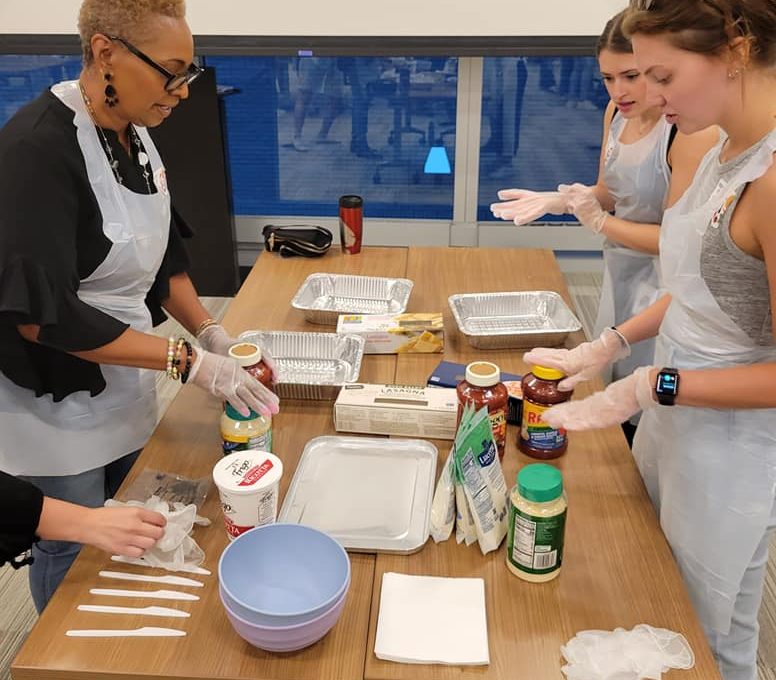 A group of four women, wearing aprons and gloves, are standing around a table during a team building cooking activity. Various ingredients, such as jars of pasta sauce, parmesan cheese, and lasagna trays, are placed on the table, along with mixing bowls and utensils. They are preparing to assemble a dish, with one woman holding a jar of sauce and the others ready to begin. The group appears focused, engaged in the process, and collaborating on the cooking task.