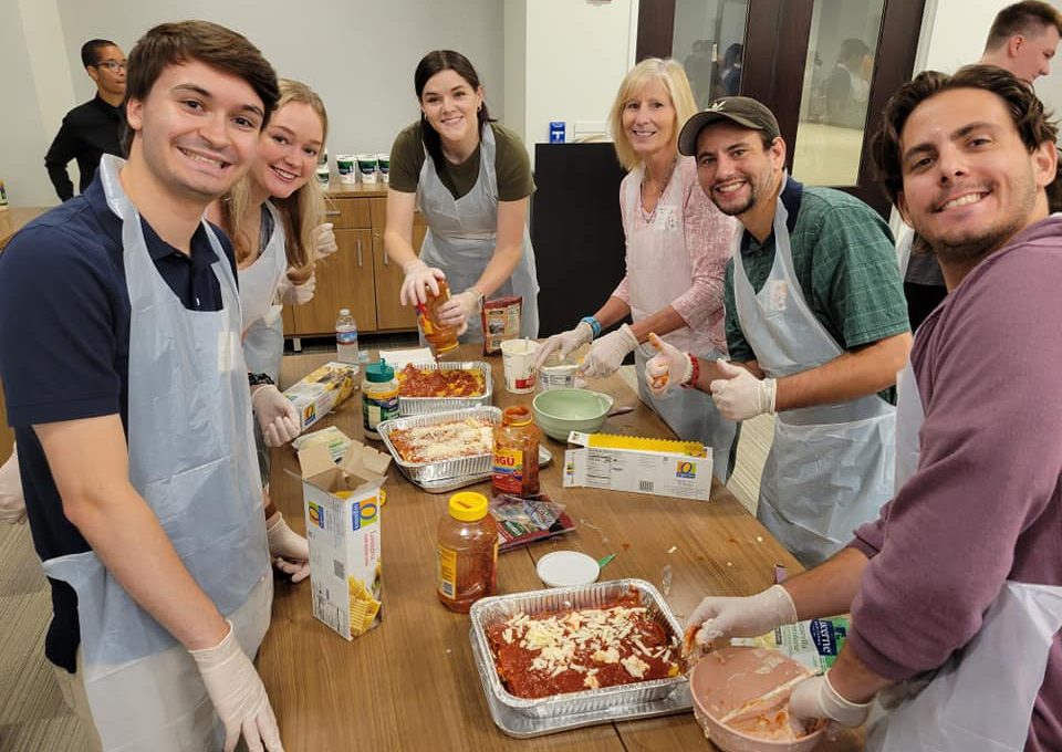 A group of six participants in the 'Cooking For A Cause' team building event smiling as they prepare meals to donate. They are gathered around a wooden table, each wearing plastic aprons and gloves while assembling dishes in aluminum trays. The team is working collaboratively, spreading sauce and layering ingredients, likely for a lasagna or similar dish. This activity combines teamwork and community service by providing meals for local food banks or soup kitchens.