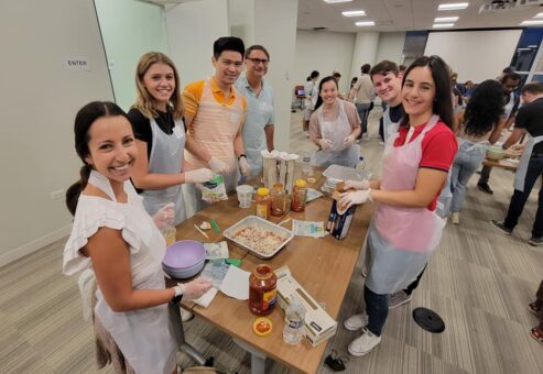 A cheerful group of seven people participating in the 'Cooking For A Cause' event. They are gathered around a wooden table, wearing plastic aprons and gloves as they work together to prepare a dish in an aluminum tray. Various ingredients like jars of sauce, cheese, and boxes of pasta are laid out on the table. This team building event focuses on creating meals to donate to local food banks or soup kitchens, combining collaboration and community service.