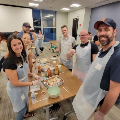 A group of six people is gathered around a table during a team building cooking activity, smiling and wearing aprons and gloves. They are preparing meals, with lasagna trays and various ingredients like pasta, sauce, and cheese laid out on the table. One member gives a thumbs-up to the camera, and the group looks cheerful and engaged in the activity. thumbnail