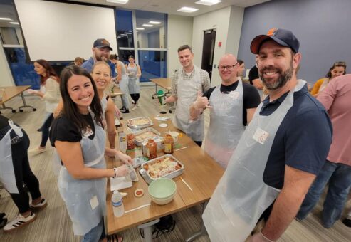 A group of six people is gathered around a table during a team building cooking activity, smiling and wearing aprons and gloves. They are preparing meals, with lasagna trays and various ingredients like pasta, sauce, and cheese laid out on the table. One member gives a thumbs-up to the camera, and the group looks cheerful and engaged in the activity.