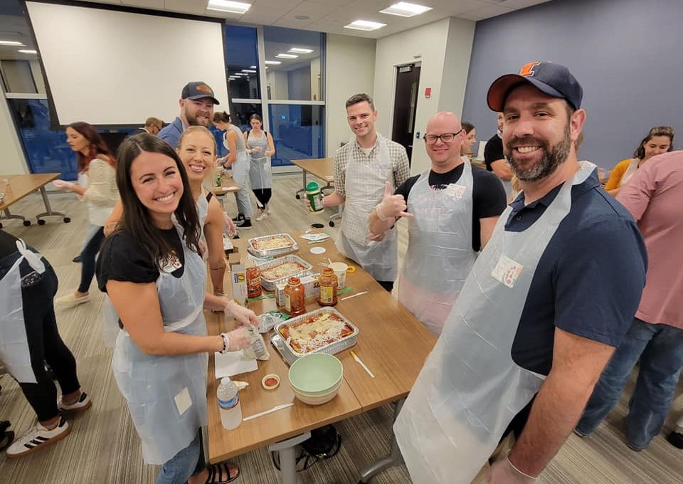 A group of six people is gathered around a table during a team building cooking activity, smiling and wearing aprons and gloves. They are preparing meals, with lasagna trays and various ingredients like pasta, sauce, and cheese laid out on the table. One member gives a thumbs-up to the camera, and the group looks cheerful and engaged in the activity.
