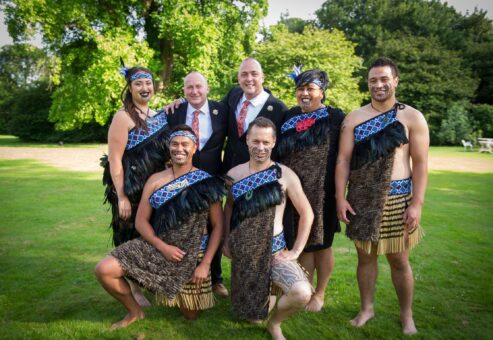 Group photo of Maori performers in traditional attire alongside two suited participants during a Haka team building event in a scenic outdoor setting.