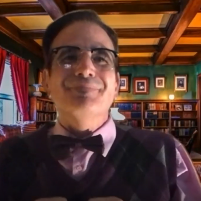 A man wearing glasses, a bow tie, and a patterned sweater vest is seated in a library setting with shelves filled with books behind him. The man is in character, part of a role-playing murder mystery team building event. thumbnail