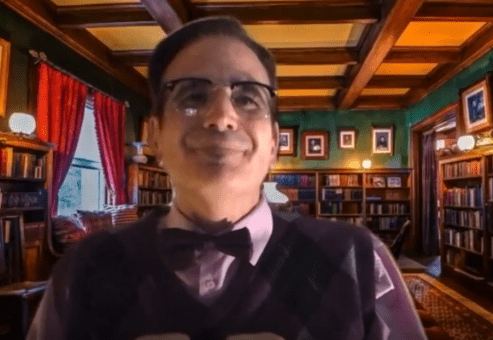 A man wearing glasses, a bow tie, and a patterned sweater vest is seated in a library setting with shelves filled with books behind him. The man is in character, part of a role-playing murder mystery team building event.