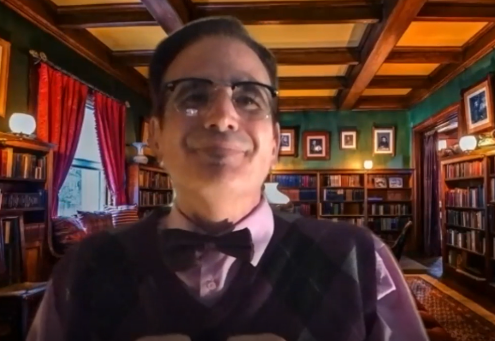 A man wearing glasses, a bow tie, and a patterned sweater vest is seated in a library setting with shelves filled with books behind him. The man is in character, part of a role-playing murder mystery team building event.