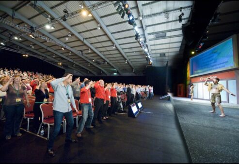 Large audience in a conference hall actively participating in a Haka team building event led by Maori performers on stage.