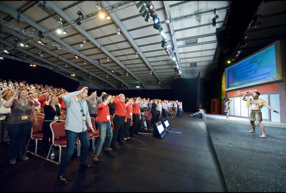 Large audience in a conference hall actively participating in a Haka team building event led by Maori performers on stage.