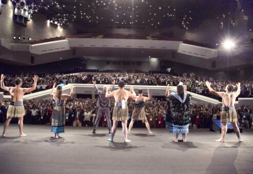Maori performers in traditional attire lead a large audience in an energetic Haka team building session on a grand stage, inspiring unity and passion.