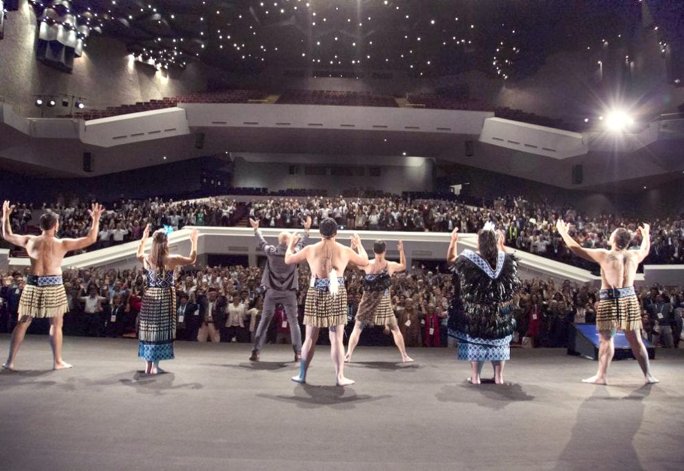 Maori performers in traditional attire lead a large audience in an energetic Haka team building session on a grand stage, inspiring unity and passion.