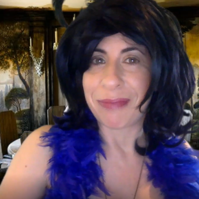 A woman with a playful expression, wearing a black wig and a bright blue feather boa, poses in front of a luxurious room decorated with an artistic mural of trees and a grand dining table. The setting evokes a sense of elegance and mystery. She is playing a role in a themed murder mystery team building event. thumbnail