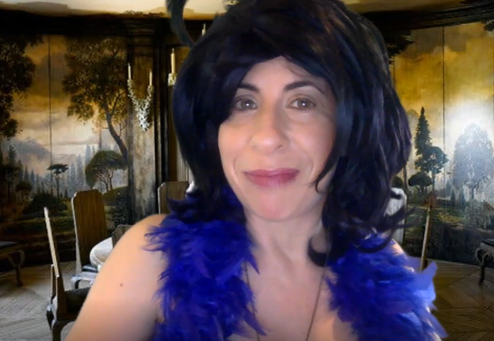 A woman with a playful expression, wearing a black wig and a bright blue feather boa, poses in front of a luxurious room decorated with an artistic mural of trees and a grand dining table. The setting evokes a sense of elegance and mystery. She is playing a role in a themed murder mystery team building event.