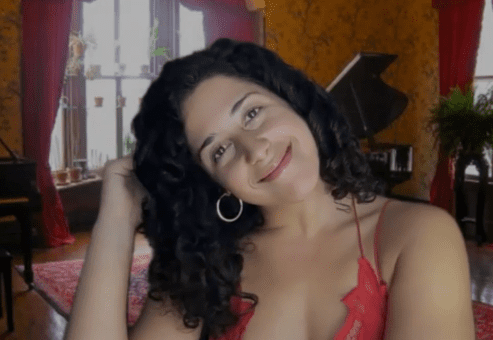 A woman with curly hair smiles while tilting her head to the side in a warm and elegantly decorated room. She is wearing a red spaghetti-strap top and hoop earrings. She's role-playing in a murder mystery team building event.