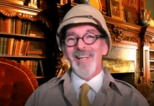 A man with glasses and a safari-style pith helmet smiles warmly in front of a cozy, vintage room filled with bookshelves. He is dressed in character, wearing a light-colored jacket and a yellow tie. The man is in character, part of a role-playing murder mystery team building event.