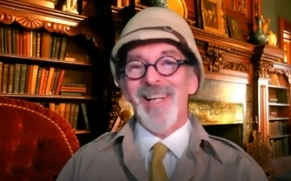 A man with glasses and a safari-style pith helmet smiles warmly in front of a cozy, vintage room filled with bookshelves. He is dressed in character, wearing a light-colored jacket and a yellow tie. The man is in character, part of a role-playing murder mystery team building event.