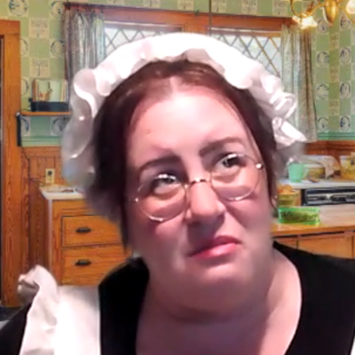 A woman dressed as a maid, wearing a white bonnet and glasses, is in an old-fashioned kitchen. She looks pensive or concerned as she gazes off to the side. The scene is part of a role-playing murder mystery. thumbnail