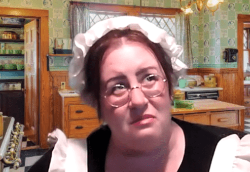 A woman dressed as a maid, wearing a white bonnet and glasses, is in an old-fashioned kitchen. She looks pensive or concerned as she gazes off to the side. The scene is part of a role-playing murder mystery.
