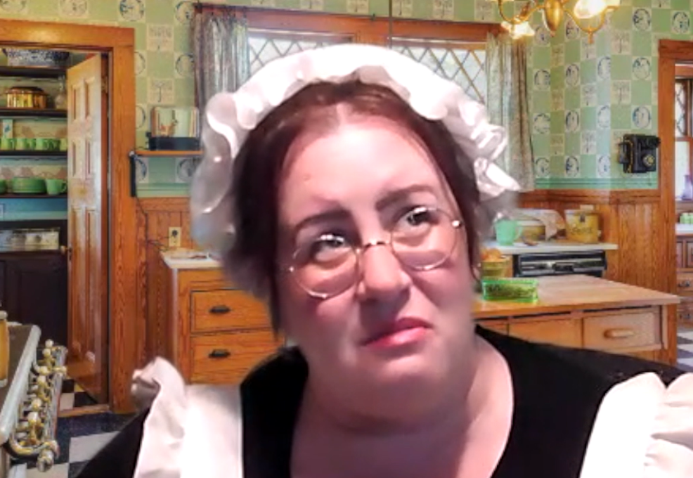 A woman dressed as a maid, wearing a white bonnet and glasses, is in an old-fashioned kitchen. She looks pensive or concerned as she gazes off to the side. The scene is part of a role-playing murder mystery.