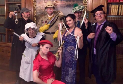 A group of seven participants pose in costume for a themed Murder Mystery Dinner team building event. Each person is dressed as a unique character, holding props that represent classic murder mystery items, such as a candlestick, rope, and revolver.