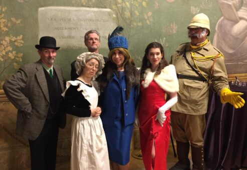 A group of actors dressed in period costumes for a Murder Mystery Dinner event. The characters include a man in a detective's coat and hat, a maid in a traditional white apron, a woman in a blue dress and fur hat, a glamorous woman in a red gown and fur shawl, and an explorer wearing a safari outfit and yellow gloves.