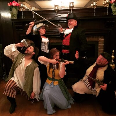 A group of actors in pirate-themed costumes pose dramatically for a Murder Mystery Dinner event. The characters include a pirate captain in a black hat and red vest, a woman dressed as a swashbuckler holding a sword, another woman in a long blue and yellow dress aiming a musket, and two additional pirates—one with a mug and the other with a flintlock pistol. They stand in front of an elegant fireplace, creating a lively and adventurous scene.
