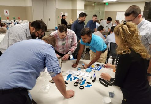 A group of participants is gathered around a table, fully engaged in assembling small robotic parts. The table is scattered with various components such as wheels, gears, and blue plastic pieces. Each person is focused on a task, contributing to the team effort to build a functioning robot during this team building event.