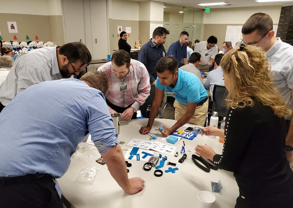 A group of participants is gathered around a table, fully engaged in assembling small robotic parts. The table is scattered with various components such as wheels, gears, and blue plastic pieces. Each person is focused on a task, contributing to the team effort to build a functioning robot during this team building event.