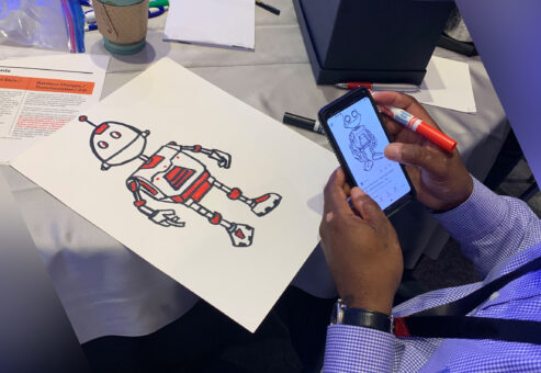A person is drawing a robot on a sheet of paper, using a reference image displayed on their smartphone. The robot has a classic, humanoid design with mechanical arms, legs, and antennae, and is being colored in with red accents. The setting appears to be part of a creative team building activity, with various supplies like markers and cups visible on the table.