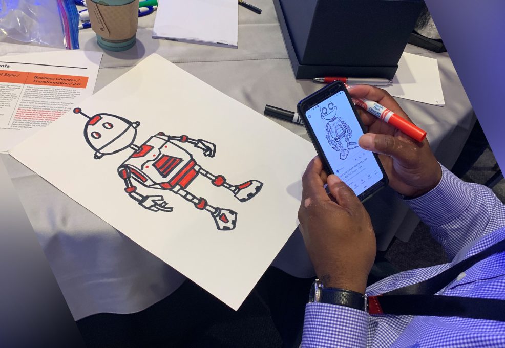A person is drawing a robot on a sheet of paper, using a reference image displayed on their smartphone. The robot has a classic, humanoid design with mechanical arms, legs, and antennae, and is being colored in with red accents. The setting appears to be part of a creative team building activity, with various supplies like markers and cups visible on the table.