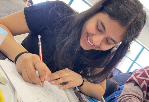 A young woman is smiling and fully engaged in drawing on graph paper. She appears focused and enjoying the creative process. She is holding a pencil and working on a sketch, with other materials spread out around her on the table.
