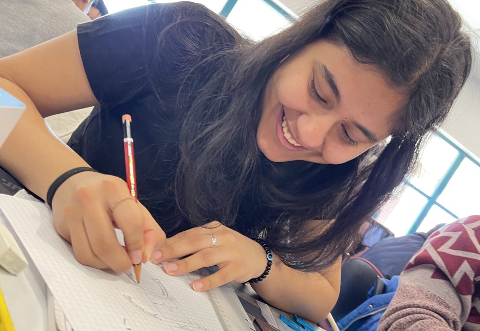 A young woman is smiling and fully engaged in drawing on graph paper. She appears focused and enjoying the creative process. She is holding a pencil and working on a sketch, with other materials spread out around her on the table.
