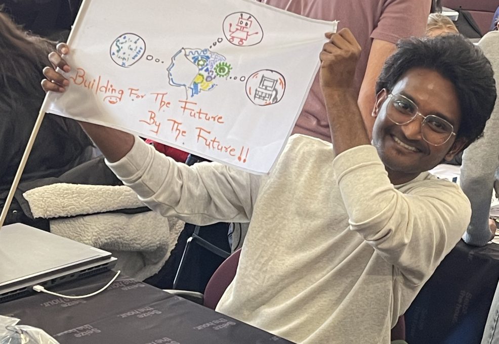 A young man is proudly holding up a flag he created, which features hand-drawn images of technology-related elements like a robot and text that reads, 