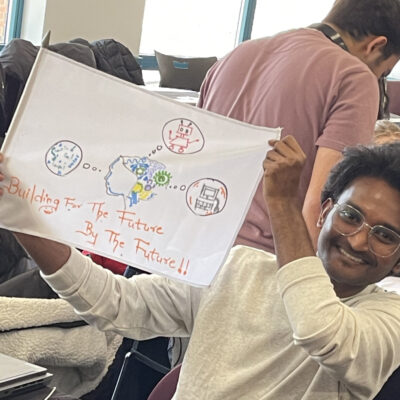 A young man is proudly holding up a flag he created, which features hand-drawn images of technology-related elements like a robot and text that reads, 