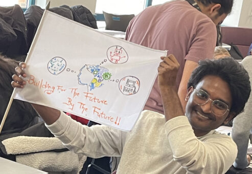 A young man is proudly holding up a flag he created, which features hand-drawn images of technology-related elements like a robot and text that reads, 
