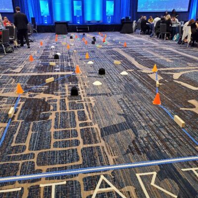 A robot obstacle course is set up indoors with orange cones, wooden blocks, and taped lines marking the track. Participants are preparing for a robot race, with 