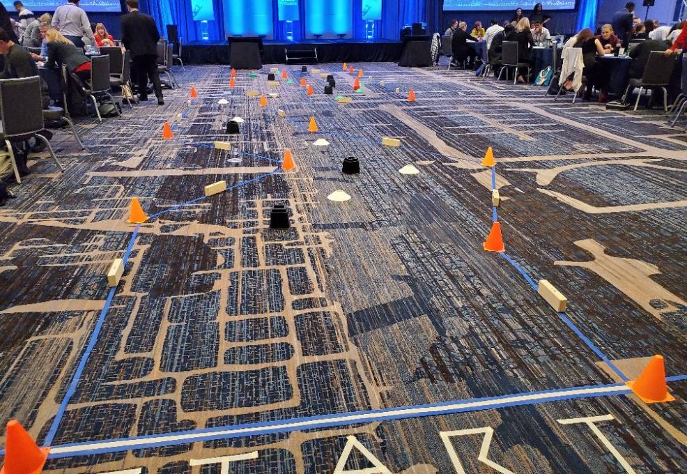 A robot obstacle course is set up indoors with orange cones, wooden blocks, and taped lines marking the track. Participants are preparing for a robot race, with 