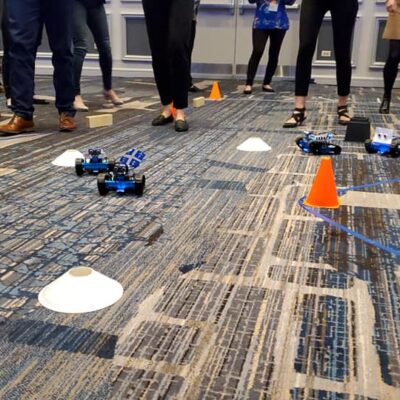 Participants are gathered around a designated race track, watching small robots they built navigate through an obstacle course marked by cones and barriers. The atmosphere is lively as teams cheer on their robots, showcasing the culmination of their team building activity. thumbnail