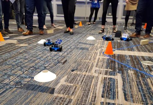 Participants are gathered around a designated race track, watching small robots they built navigate through an obstacle course marked by cones and barriers. The atmosphere is lively as teams cheer on their robots, showcasing the culmination of their team building activity.