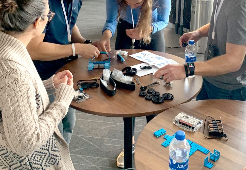 A group of people collaborate on building a robot at a table, using various robot parts, instructions, and tools during a robotics team building event. Bottled water and a battery kit are also on the table.