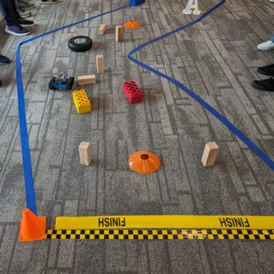 A small robot is navigating through an obstacle course marked by blue tape on the floor. Various objects, such as large toy bricks, wooden blocks, and traffic cones, create the course's challenges. The 