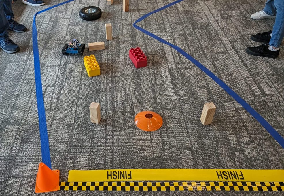 A small robot is navigating through an obstacle course marked by blue tape on the floor. Various objects, such as large toy bricks, wooden blocks, and traffic cones, create the course's challenges. The 