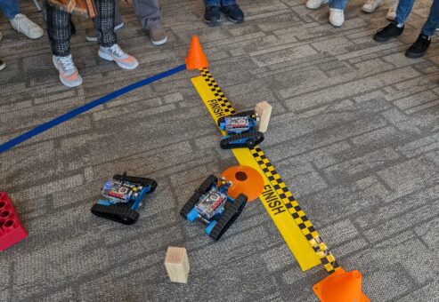 Three small robots are racing across a finish line in a friendly competition. Participants are gathered around, watching the robots as they navigate obstacles on the course. This image represents the final stage of a robotics team building event.