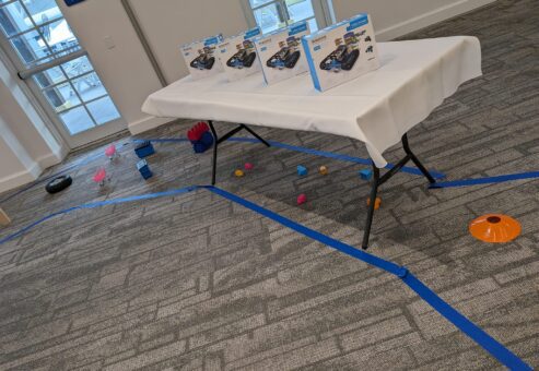 A table is set up with robot kits ready for assembly, placed in a room where a robotics team building event is taking place. Blue tape marks the floor for an obstacle course, alongside cones and other game objects.