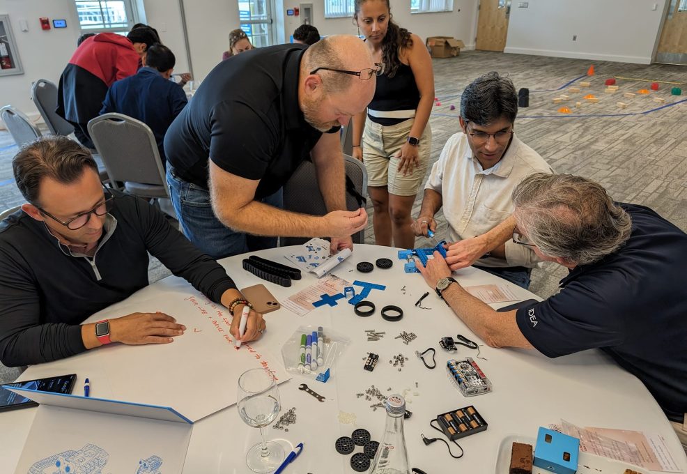 A group of participants is working together to assemble a robot. The table is filled with parts, tools, and instructions as they collaborate during a robotics team building event.
