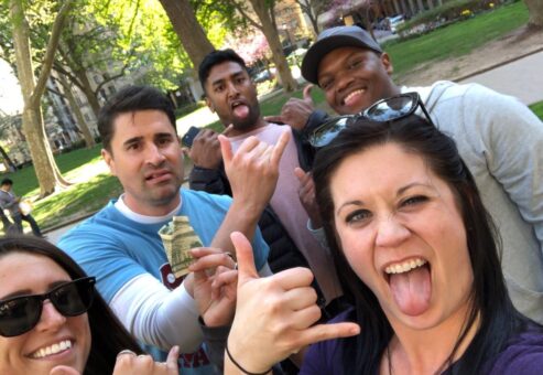 Team making funny faces and gestures, holding Monopoly money during a playful moment in the Team-opoly scavenger hunt event outdoors.