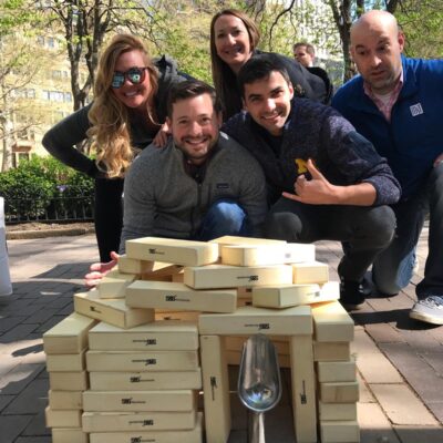 Team proudly posing behind a structure made from oversized wooden blocks during a challenge in the Team-opoly scavenger hunt event. thumbnail