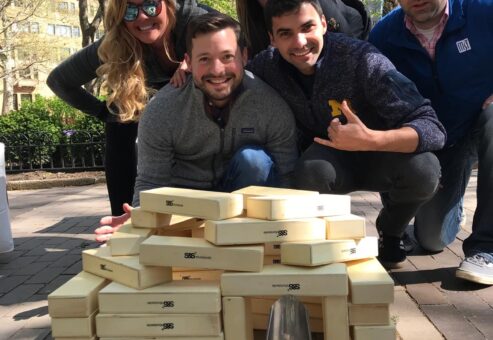 Team proudly posing behind a structure made from oversized wooden blocks during a challenge in the Team-opoly scavenger hunt event.