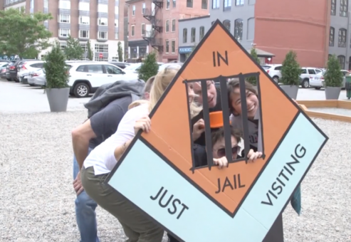 Group posing with a life-sized 'In Jail' game piece during a Team-opoly scavenger hunt team building event.